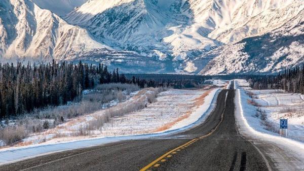 A paved road with yellow lines stretches toward a tall, snow covered mountain
