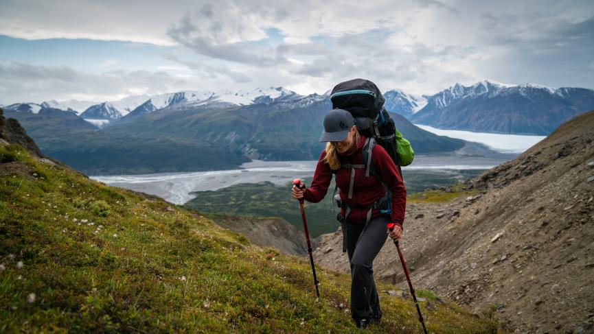 A person in a red hiking sweater and a large backpack uses hiking poles to climb the side of a mountain.
