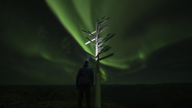 A person stands beside a white sign post under the aurora