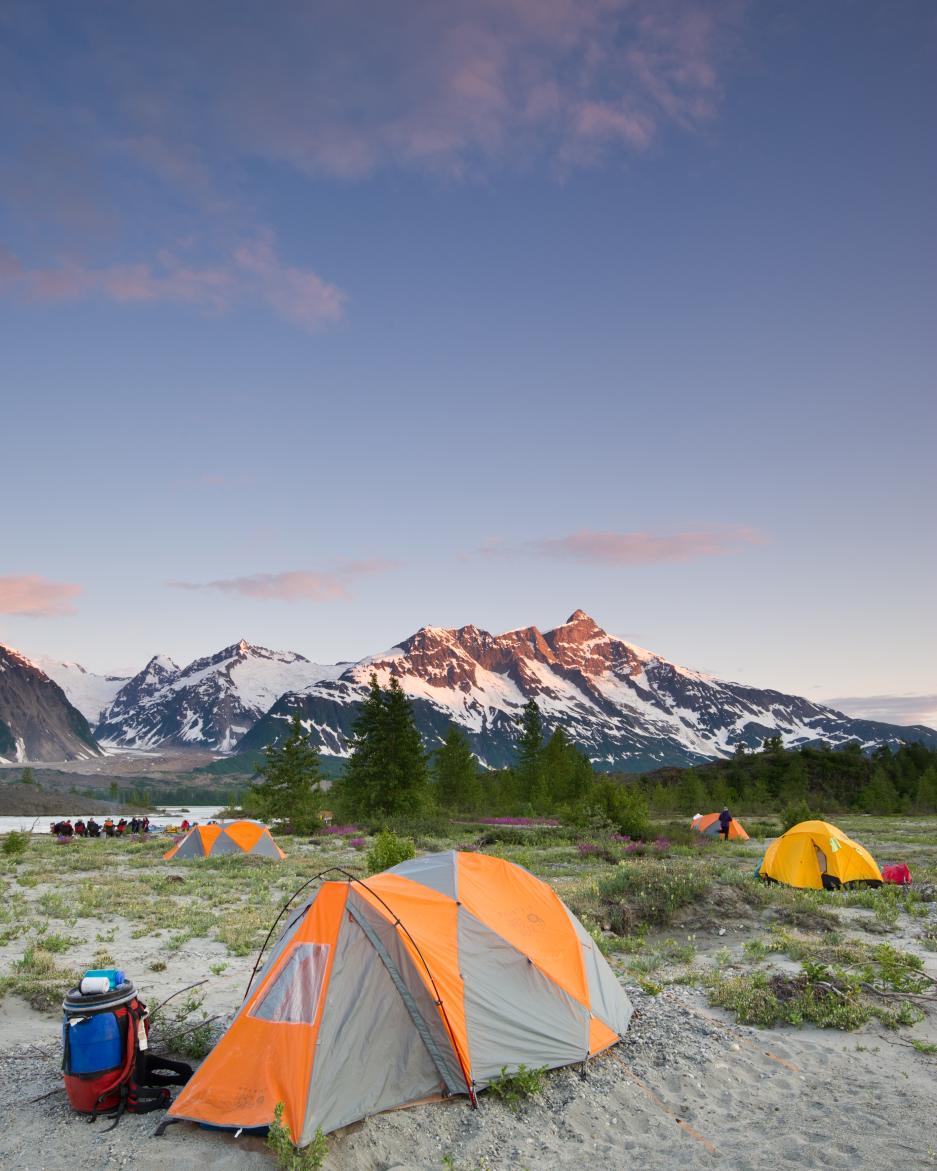 An orange and grey tent is pitched on the sand by a river under a bright blue sky. There is a snowy mountain in the background.